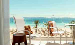 Näkymä parvekkeelta rannalle, jossa on aurinkotuoleja ja -varjoja, palmuja sekä kauempana avomeri ja vuoristo. (Tekoälyn luoma teksti)