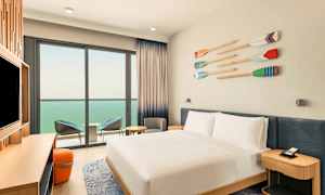 Hotellihuone, jossa on valkoiseksi pedattu parivuode, televisio tasolla ja istuinryhmä parvekkeen vieressä, jossa on näkymä merelle. (Tekoälyn luoma teksti)