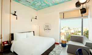 Hotellihuone, jossa suuri sänky, moderni sisustus ja värikäs katto. Suuresta ikkunasta avautuu näkymä kaupunkiin. (Tekoälyn luoma teksti)