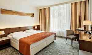 Hotellihuone, jossa on parivuode, oranssi päiväpeitto ja kaksi valkoista tyynyä. Molemmin puolin on yöpöydät lampuilla, ja taustalla näkyy suuri ikkuna verhoineen. Oikealla puolella on kirjoituspöytä tuolin ja erilaisten esineiden kanssa. (Tekoälyn luoma teksti)