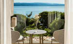 Pöytäkoriste, jossa kukkia, pöydän ääressä ikkunan edessä, näkymä merelle ja vehreä luonto. (Tekoälyn luoma teksti)