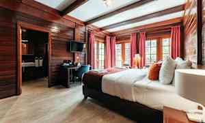 Hotellihuone, jossa puusisustus, leveä sänky valkoisilla ja värikkäillä tyynyillä, punaiset verhot ja ikkunat näkymällä. (Tekoälyn luoma teksti)