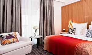 Hotellihuone, jossa sänky, kuvioitu tyyny, punainen viltti, tuoli ja pöytä, jossa kukkia. (Tekoälyn luoma teksti)