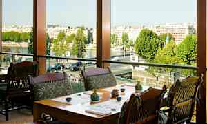 Ravintola, jossa on pöytiä ja tuoleja suuren ikkunan lähellä, näkymä kaupunkiin ja joelle. (Tekoälyn luoma teksti)