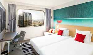 Hotellihuone, jossa on kaksi yhden hengen vuodetta, työtila ja suuri ikkunanäkymä kaupungin siluettiin. (Tekoälyn luoma teksti)