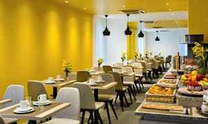 Moderni ravintola-alue, jossa on keltaiset seinät, katettuja pöytiä ja seisova pöytä erilaisine ruokalajeineen. (Tekoälyn luoma teksti)