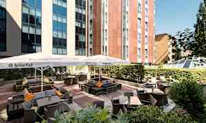 Kuva ulkoilmaravintolasta, jossa on pöytiä, tuoleja ja aurinkovarjoja rakennuksen vieressä. (Tekoälyn luoma teksti)