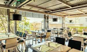 Ravintola, jossa on useita katettuja pöytiä ja tuoleja, ikkunoista kaupunkinäkymä. (Tekoälyn luoma teksti)