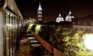Kattoalue, jolla on pöytiä ja tuoleja, ympäröitynä vihreillä kasveilla, kirkon kupolit ja kellotorni näkyvät taustalla illalla. (Tekoälyn luoma teksti)