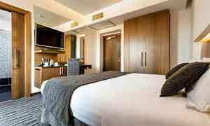 Hotellihuone, jossa on suuri sänky, kirjoituspöytä tuoleineen ja seinälle kiinnitetty televisio sekä puukaapit. (Tekoälyn luoma teksti)