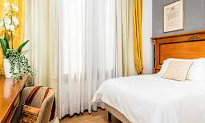 Hotellihuone, jossa on sänky, kirjoituspöytä ja tuoli. Ikkunan edessä on verhot. (Tekoälyn luoma teksti)