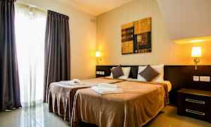 Hotellihuone, jossa on kaksi yhden hengen sänkyä, yöpöydät, seinävalaisimet, maalaus sänkyjen yläpuolella ja verhot ikkunan edessä. (Tekoälyn luoma teksti)