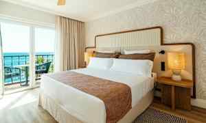 Hotellihuone, jossa on iso sänky, kaksi yöpöytää lampuilla ja parveke merinäköalalla. (Tekoälyn luoma teksti)