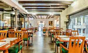 Ravintolan sisätila, jossa on tyhjiä pöytiä ja tuoleja, puupalkkeja katossa ja suuret ikkunat kadulle päin. (Tekoälyn luoma teksti)