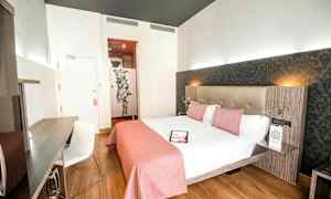 Valoisa hotellihuone, jossa on parisänky valkoisilla lakanoilla ja vaaleanpunaisilla vuodevaatteilla, kirjoituspöytä tuoleineen ja koristeellinen seinä sängyn takana. (Tekoälyn luoma teksti)