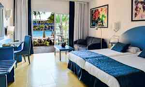 Hotellihuone, jossa on iso sänky, oleskelualue ja näkymä uima-altaalle lasiovien kautta. (Tekoälyn luoma teksti)