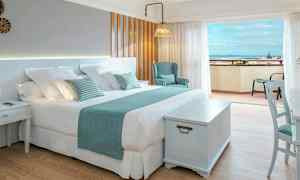 Vaalea hotellihuone, jossa suuri parivuode, tuoli ja pöytä, sekä parveke merinäköalalla. (Tekoälyn luoma teksti)