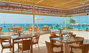 Kuva ravintolasta, jossa on punottuja tuoleja ja lasipöytiä. Sijainti meren äärellä, näkymä siniselle vesihorisontille. (Tekoälyn luoma teksti)