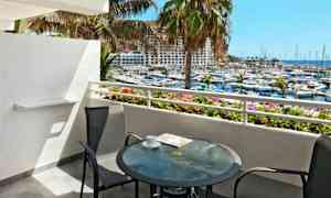 Näkymä parvekkeelta, jossa lasipöytä ja kaksi tuolia, jotka osoittavat kohti satamaa ja merta. (Tekoälyn luoma teksti)