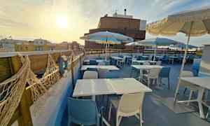 Kuvassa näkyy terassi, jossa on pöytiä, sinisiä ja valkoisia tuoleja sekä aurinkovarjoja aurinkoisella taivaalla. (Tekoälyn luoma teksti)