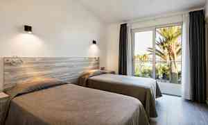 Kaksi yhden hengen sänkyä valoisassa huoneessa, jossa on suuret ikkunat parvekkeelle ja näkymä palmuihin. (Tekoälyn luoma teksti)