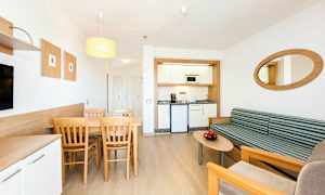 Moderni hotellihuone, jossa on pieni keittiönurkkaus, ruokailutila pöytineen ja tuoleineen sekä oleskelutila sohvan ja sohvapöydän kanssa. (Tekoälyn luoma teksti)