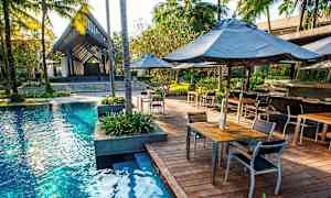 Ulkoallas, jota ympäröi puinen terassi, jossa on pöytiä ja tuoleja. Palmuja ja trooppisia kasveja ympäröi alue. (Tekoälyn luoma teksti)