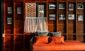 Huone, jossa on puuseinät, koristeltuna mustavalkoisilla valokuvilla ja oranssi istuin pehmeillä tyynyillä ikkunan vieressä pitsiverhon kanssa. (Tekoälyn luoma teksti)