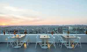 Kattoterassi, jolla on pöytiä ja tuoleja sekä näkymä kaupungin yli auringonlaskun aikaan. (Tekoälyn luoma teksti)