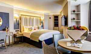 Valoisa hotellihuone, jossa on parivuode, tuoleja ja hedelmäkulho. (Tekoälyn luoma teksti)