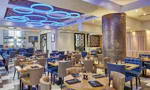 Ravintola, jossa on pöytiä ja tuoleja, moderni sisustus ja koristeellinen valaistus. (Tekoälyn luoma teksti)