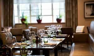 Ravintola-alue, jossa on katetut pöydät, laseja ja lautasliinoja. Taustalla ikkunoita, joissa on kasveja. (Tekoälyn luoma teksti)