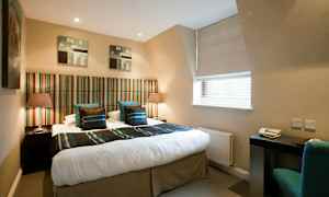 Hotellihuone, jossa on parivuode, raidallinen sängynpääty ja kaksi yöpöytää lampuilla. Kirjoituspöytä ja tuoli ikkunan ääressä. (Tekoälyn luoma teksti)