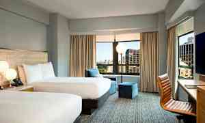 Hotellihuone, jossa on kaksi parisänkyä, suuret ikkunat kaupunkinäkymällä, nojatuoli ja työpöytä tuolilla. (Tekoälyn luoma teksti)