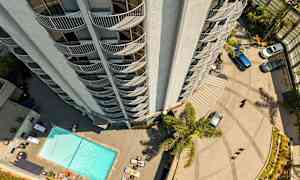 Korkea rakennus kuvattuna ylhäältä, ja sen ulkonevat parvekkeet erottuvat. Alapuolella on uima-allas, aurinkotuoleja ja pysäköityjä autoja. (Tekoälyn luoma teksti)