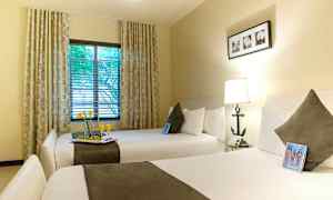 Hotellihuone, jossa on kaksi leveää vuodetta, kummallakin tyyny ja peite. Ikkuna, jossa verhot ja näkymä puille. (Tekoälyn luoma teksti)