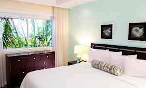 Makuuhuone, jossa on sänky, kaksi taideteosta, ikkunasta näkyy vehreitä palmuja ja lipasto. (Tekoälyn luoma teksti)