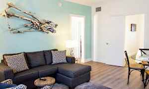 Moderni olohuone, jossa on harmaa kulmasohva, lasipöytä ja istuinalue kuvioiduilla tyynyillä. Vaaleansinisellä seinällä on koristeellinen metalliveistos. (Tekoälyn luoma teksti)