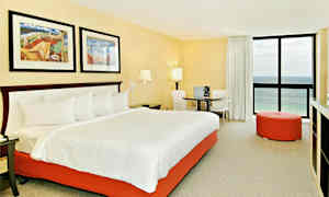 Hotellihuone, jossa on suuri sänky valkoisilla vuodevaatteilla ja oranssi sängyn pohja. Huoneessa on kaksi taulua sängyn yläpuolella, pyöreä pöytä ja kaksi tuolia, oranssi istuinpuffi ja ikkuna, josta avautuu näkymä merelle. (Tekoälyn luoma teksti)