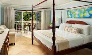 Huoneessa on neljä pylvässänky ja vaaleat kalusteet. Parveke vehreine palmujenäkymineen näkyy avoimien lasiovien läpi. (Tekoälyn luoma teksti)