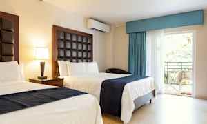 Hotellihuone, jossa kaksi sänkyä, vaaleat seinät ja tummat sängynpäädyt. Pöytä lampulla ja parvekkeen ovi näkymällä vehreyteen. (Tekoälyn luoma teksti)