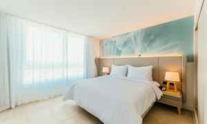 Valoisa ja modernisti sisustettu makuuhuone, jossa parivuode, kaksi yöpöytää ja suuri ikkunapaneeli ohuilla valkoisilla verhoilla. (Tekoälyn luoma teksti)