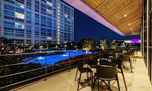 Näkymä valaistulle uima-altaalle terassilta, jossa on pöytiä ja tuoleja, ympäröity kasvillisuudella ja rakennus taustalla illallisaikaan. (Tekoälyn luoma teksti)