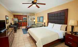 Hotellihuone, jossa on iso sänky, yöpöydät, verhoiltu nojatuoli, ruokailutila ja keittiö taustalla. (Tekoälyn luoma teksti)