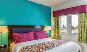 Makuuhuone, jossa on parivuode, värikkäitä vuodevaatteita ja tyynyjä. Seinät ovat turkooseja, ja huoneesta on näkymä palmuihin ja merelle. (Tekoälyn luoma teksti)