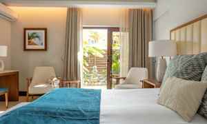 Valoisa ja moderni makuuhuone, jossa on sänky valkoisilla ja sinisillä vuodevaatteilla, kaksi tuolia ja pieni pöytä lasioven ääressä näkymällä puutarhaan. (Tekoälyn luoma teksti)