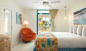 Makuuhuone, jossa on vaalea sisustus. Vuode, jonka päällä on värikäs päiväpeite, oranssi nojatuoli, pöytälamppu ja ikkuna, josta avautuu näkymä ulos. (Tekoälyn luoma teksti)