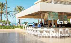 Ulkobaari katoksella ja korkeilla tuoleilla, hiekkarannan lähellä, palmuja ja sininen taivas taustalla. (Tekoälyn luoma teksti)