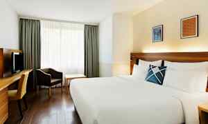 Hotellihuone, jossa suuri sänky valkoisine vuodevaatteineen, työpöytä tuoleineen, nojatuoli ja pöytä ikkunan edessä verhoineen. (Tekoälyn luoma teksti)