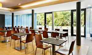 Valoisa ruokailutila, jossa on pöytiä ja tuoleja suurten ikkunoiden ääressä ja näkymä vihreisiin pensaisiin. (Tekoälyn luoma teksti)